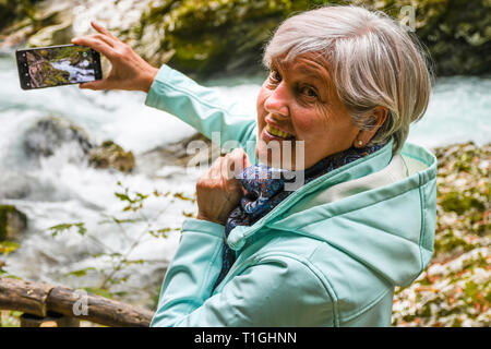 Schöne attraktive ältere, reife Frau mit glänzenden grauen Haaren fotografieren und selfies Outdoor in einer Schlucht Vintgar River Canyon Schlucht an einem sonnigen Tag Stockfoto