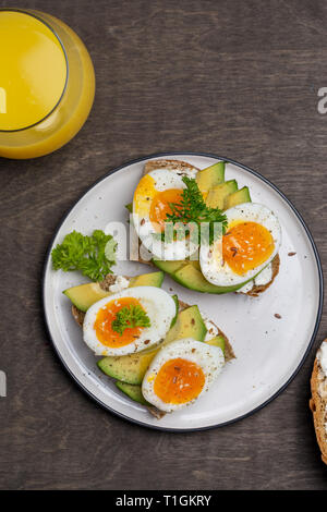 Zwei leckere Sandwiches mit Avocado und gekochtem Ei auf weiße Platte, Vollkornbrot mit Frischkäse und Glas Orangensaft in der Nähe von auf hölzernen Hintergrund Stockfoto