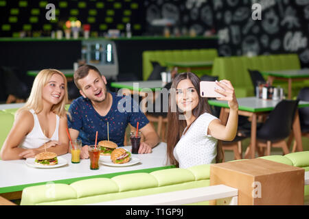 Firma glücklich lächelnd, am Telefon und unter selfie beim Essen leckere Burger im Cafe. Freunde Rest in und ejoying gemeinsam entspannen. Konzept von Freundschaft und Glück. Stockfoto