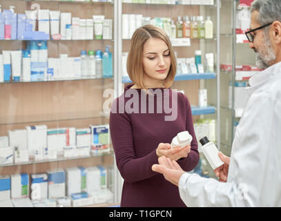 Weibliche Kunden im Gespräch mit dem Apotheker in der Apotheke. Apotheke Spezialisten helfen Client mit kosmetischen Produkten. Frau und Mann im weißen Mantel mit weißem kosmetische Flaschen. Stockfoto