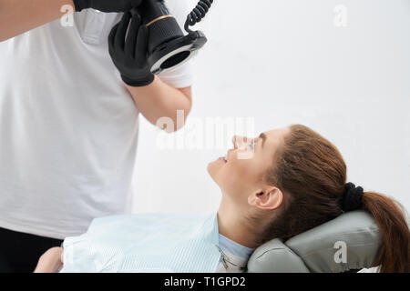 Seitenansicht der jungen Patientin sitzen in Zahnarztpraxis und schönes Lächeln nach dem Verfahren. Arzt unter Foto von Ergebnis nach dem Bleichen der Zähne. Konzept der Zahnheilkunde. Stockfoto
