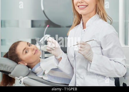 Zahnarzt, Arzt und Patient posing und lächelnd in der Zahnheilkunde Klinik. Schöne stomatologist tragen in weiße Uniform, Handschuhe holding Restaurierung Instrumente. Kunden liegen auf zahnarztstuhl. Stockfoto