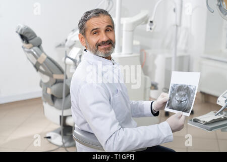 Happy Zahnarzt in der Zahnheilkunde Klinik arbeiten. Gut aussehender bärtiger Mann mit zahnmedizinischen Zähne scannen. Reifen Spezialist für weiße Uniform mit Blick auf die Kamera oder Lächeln und posieren. Stockfoto