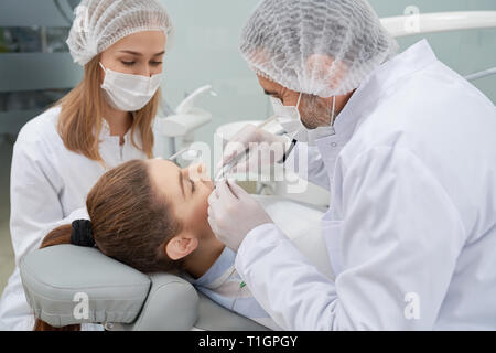 Professionelle Arzt und Helfer arbeiten mit Kunden in der Zahnheilkunde Klinik. Liegende Frau auf Zahnarztstuhl, Zähne getan. Stomatologist mit speziellen Werkzeugen und Geräten. Stockfoto