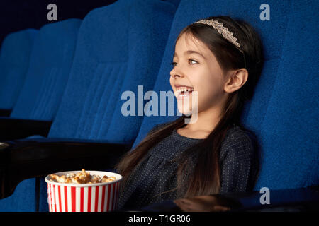 Schöne, hübsche kleine Mädchen watchong Film in Kino, sitzen in Dunkelblau bequeme Stühle, holding Popcornwanne. Fröhliches Kind aufzupassen Film, lachen, genießen Film Premiere. Stockfoto