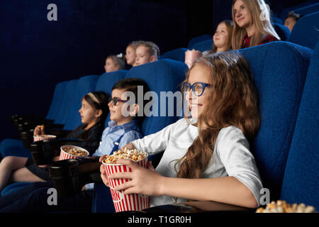 Glückliche, fröhliche Kinder sitzen in Dunkelblau, bequeme Stühle im modernen Kino Theater, Film oder Cartoon. Kinder genießen Film, lächelnd, holding Eimer mit Popcorn. Stockfoto