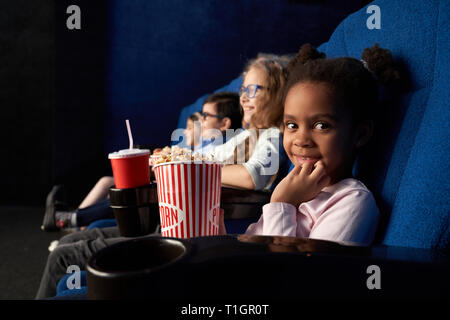 Kleine, niedliche afrikanisches Mädchen im Kino sitzen mit Freunden, holding Popcornwanne, an der Kamera schaut. Kinder Premiere von Cartoon oder Film im Kino genießen. Stockfoto