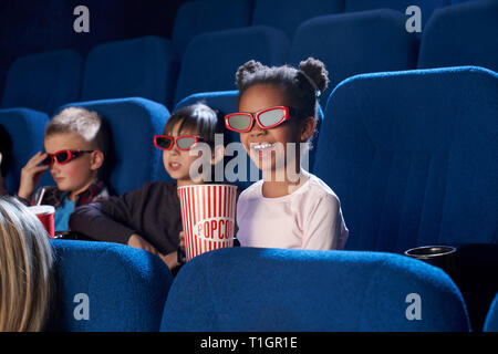 Freundliche, fröhliche Kinder im Kino sitzen, Film oder Cartoon. Kinder tragen in 3-D-Brille. Süße kleine Mädchen lächelnd, holding Popcornwanne. Stockfoto