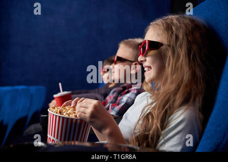 Seitenansicht des schönen Mädchen tragen in 3D-Brille, holding Popcornwanne, Lächeln, genießen Film Premiere. Kinder im Kino sitzen, beobachten aufmerksam Film oder Cartoon. Stockfoto