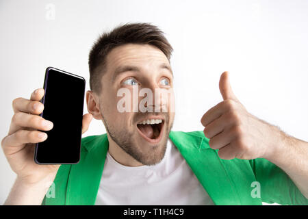 Sein kühles. Gute Nachrichten. Tun, wie mich. Jungen gutaussehenden Mann zeigt der Bildschirm des Smartphones und Unterzeichnung OK Zeichen auf grauem Hintergrund. Menschliche Emotionen, Fa Stockfoto