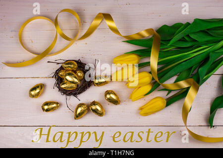 Ostern Hintergrund. Ostern goldene Eier und gelbe Tulpen. Auf einem weißen Holz- Hintergrund. Aufschrift Frohe Ostern. Platz für Text Stockfoto