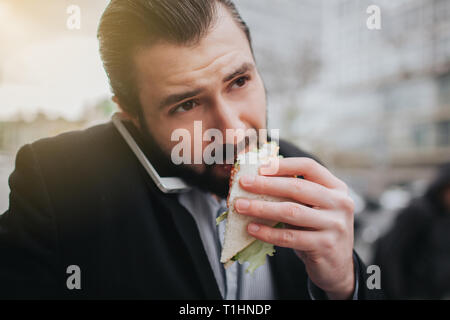 Viel beschäftigter Mann ist in Eile, hat er keine Zeit, er wird Snack auf der Essen zu gehen. Arbeitnehmer essen, trinken Kaffee, telefonieren, gleichzeitig Stockfoto