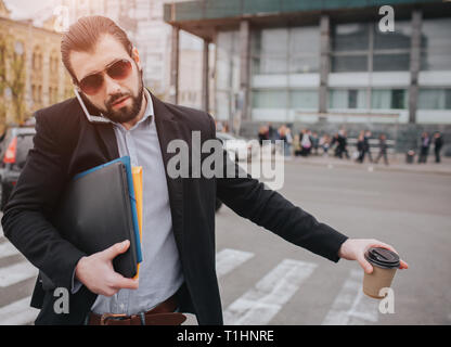 Geschäftsmann mit Kaffee Tasse man ein Taxi Bärtigen. Er hat keine Zeit, er wird am Telefon zu sprechen. Mann da mehrere Aufgaben Stockfoto
