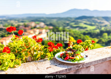 Rucola Salat mit Oliven auf Teller auf Balkon Terrasse mit roten Geranien Blumen im Garten draußen in Italien in der Toskana Bergblick Stockfoto