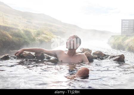 Junge glücklicher Mann Baden in heißen Quellen in Hveragerdi Reykjadalur im Herbst morgen Tag im Süden Islands auf golden circle Felsen und Fluss Dampf Stockfoto