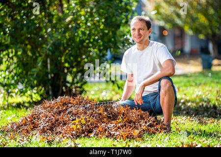 Junger Mann Hausbesitzer Sitzen im Garten Garten Garten harken trockenen Herbst Laub Eichenlaub Hausbesetzung mit Rechen im sonnigen Herbst Stockfoto