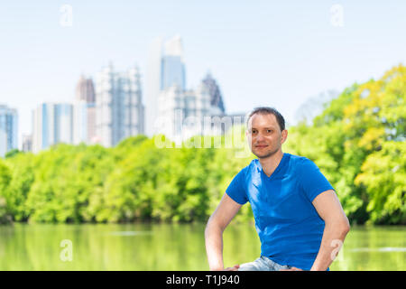 Junge glücklich Mann auf Geländer in Piedmont Park in Atlanta, Georgia sitzt lächelnd mit malerischem Stadtbild Skyline der Stadt Wolkenkratzer der Innenstadt in sp Stockfoto