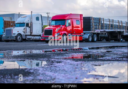 Verschiedene Motorhaube Marken und Modelle der professionellen big Rigs semi Trucks mit kommerziellen Ladung auf Auflieger in Reihe stehen auf der industriellen Parkin Stockfoto