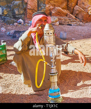 DAHAB, Ägypten - Dezember 16, 2017: Das Porträt der älteren Beduinen, Shisha rauchen, sitzen auf den Sand im Schatten der roten Felsen des Kleinen Farbe Stockfoto