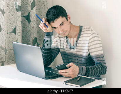 Portrait des jungen Mannes in Freizeitkleidung auf dem Job Arbeiten am Laptop, Smartphone und mit nachdenklichen Gesicht Stockfoto