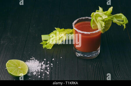 Cocktail Bloody Mary mit Kalk und Sellerie, Salz und Snacks in einem Glas auf einem schwarzen Holztisch. Stockfoto