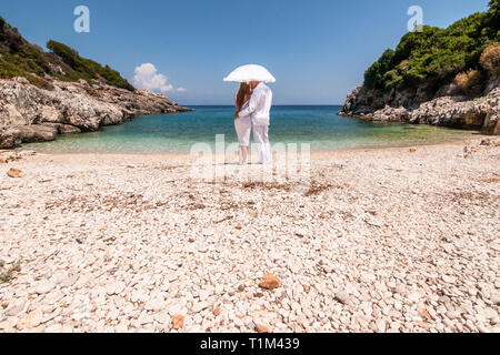 Romantische Ansicht der Paare in weißen Kleidern kuscheln unter weißen Regenschirm, stehend auf felsigen Strand und Blick auf die malerische Landschaft der blauen Meer. Anhand von quantitativen Simulatio Stockfoto