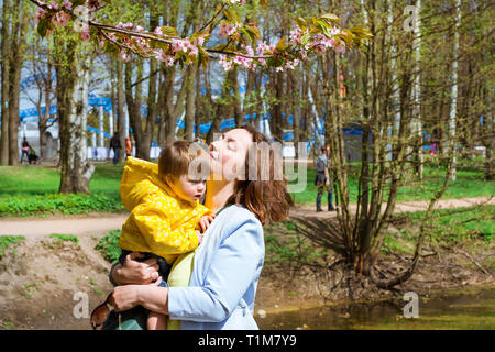 Junge Frau mit einem Kind auf dem Arm zufrieden Sakura Blüten im Frühling Park. Freude an der Mutterschaft und die Entwicklung des Kindes. Kirschblüte Baum im späten Frühjahr. Flora im Nordwesten von Russland, St. Petersburg Stockfoto