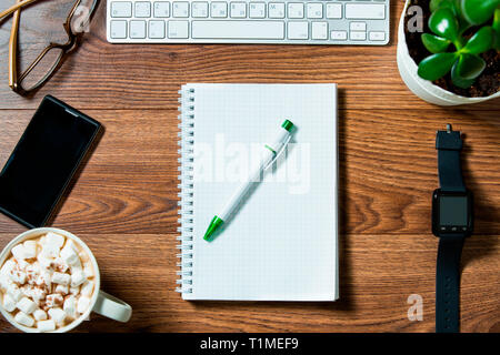 Business Workplace von oben, Flay Lay. Tastatur, Smart Watch, Smartphone, Brille, Papier-Notizbuch, heiße Schokolade mit Marshmallow auf dem dunklen Holz Stockfoto