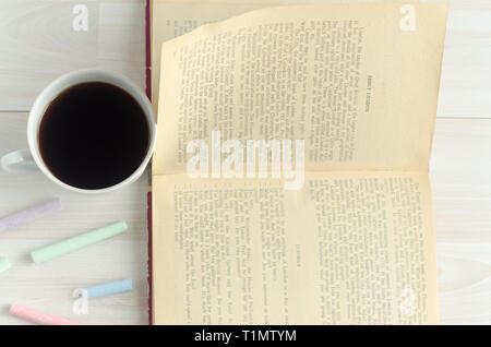 Eine Tasse Kaffee, bunte Kreide und ein offenes Buch auf weißem Holz- Hintergrund. Welttag des Buches, flach. Stockfoto