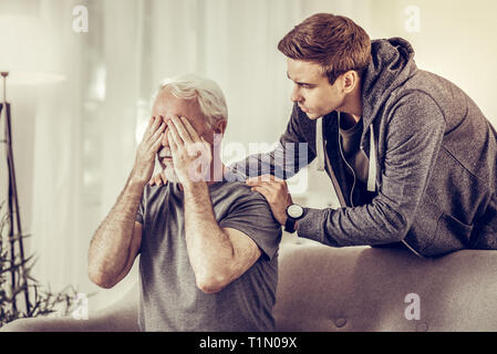 Junge attraktive umgekippt Mann calmingly umarmen Kranke schmerzhaften grauhaarige Großvater Stockfoto