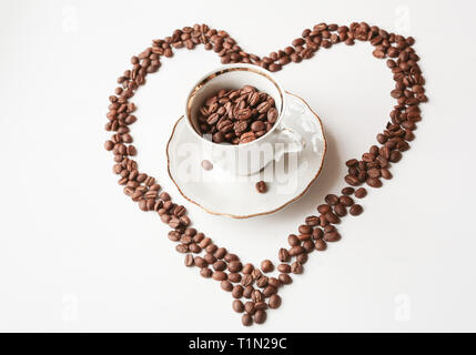 Kaffeebohnen im weißen Porzellan Schale auf die weiße Platte, surraunded der Kaffeebohnen, die wie ein Herz gelegt wird Stockfoto