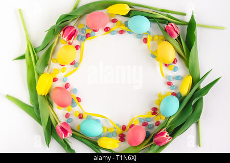 Buntes Osterfest Kranz aus gelben Tulpen, bunte Eier und Süßigkeiten auf weißem Hintergrund. Flach. Stockfoto