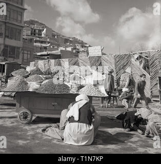 1960er Jahre, Afghanistan, Straße in Kabul, zeigt einen Afghane in traditioneller Kleidung, der an seinem fahrbaren Wagen sitzt und die Lebensmittel, die er verkauft, Stapel getrockneter Nüsse und Früchte, Heftklammern der afghanischen Ernährung zeigt. Stockfoto