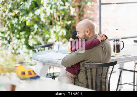 Liebevoller Vater und Sohn umarmen am Tisch Stockfoto