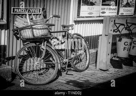 Monochrome Nahaufnahme des alten britischen Vintage-Fahrrad, Fahrrad, Korb auf der Vorderseite gegen Paketbüro auf Plattform des britischen Heritage Bahnhof ruht. Stockfoto