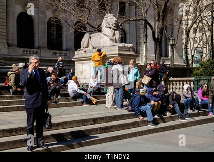 Ein Geschäftsmann im Anzug mit Aktenkoffer Gespräche über sein Handy vor der New York Public Library an der 5th Ave. Menschen auf der Treppe hinter sitzen. Mar 2018 Stockfoto