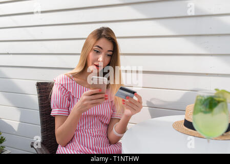 Überrascht Frau per Rechnung, per Telefon. Das Mädchen im Sommer Cafe auf der Veranda des Restaurants. Sitzt an einem Tisch in der Hand smartphone, online banking online Stockfoto