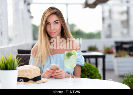 Schöne Mädchen mit langen Haaren. Eine Frau Sommer Cafe Restaurant. Sitzt am Tisch blaues Kleid, close-up. In hand, Telefon, Tisch Glas cocktail Stockfoto