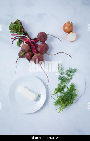 Blick von oben auf die rohe rote Beete, Ziegenkäse auf eine weiße Platte, Zwiebel halbieren und Dill. Gesunde vegetarische Zutaten auf einem hellen Marmorboden Oberfläche. Stockfoto