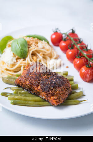 Nahaufnahme der geschwärzten Kabeljau mit grünem Gemüse, Tomaten, Nudeln in eine weiße Platte mit einem weißen Hintergrund. Gebratene gewürzten Fisch. Stockfoto