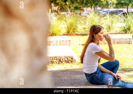 Eine schöne junge Frau in einem weißen Hemd Setzen auf ihre Sonnenbrille beim Sitzen auf einem Betonklotz an einem sonnigen Tag. Stockfoto