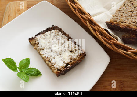 Braunes Brot mit Butter auf weiße Platte mit grünem Basilikum, andere Schichten in einem Korb, Landhausstil. Stockfoto