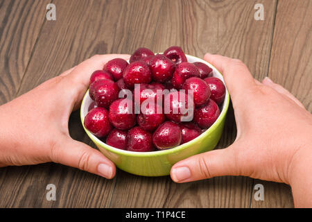 Süßkirschen Flor. Grüne Schüssel. Die menschliche Hand. Holz Tisch. Prunus avium. Frisch feucht rote Früchte gepflückt. Reife saftige Beeren in Keramik Teller. Wassertropfen. Stockfoto