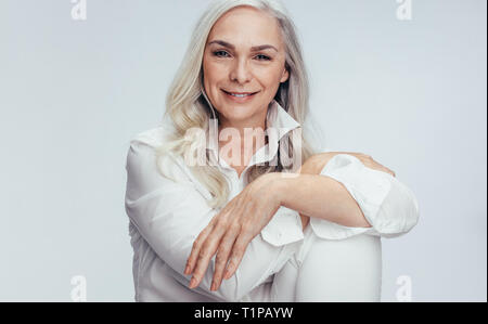 Wunderschöne reife Frau über weißen Hintergrund sitzen. Ältere Frau sitzt locker im Studio. Stockfoto