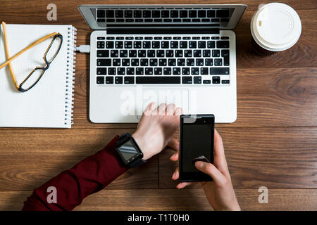 Business Person im Büro Schreibtisch arbeiten, schreiben und mit Laptop, Ansicht von oben. Smart Watch an Hand und Smart Phone auf dem Tisch. Kaffeetasse, Notepad Stockfoto
