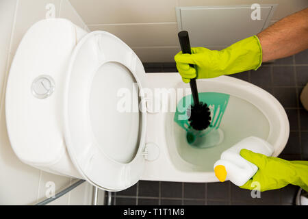 Mann in Gummi Schutzhandschuhe reinigen, waschen Wc mit Pinsel und Flasche Reinigungsmittel in Händen. Grüne Flüssigkeit tropft in der WC-Reiniger Stockfoto