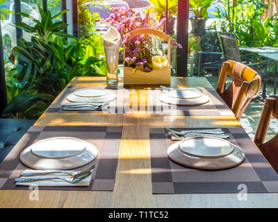 Weiße Platten, Edelstahl, Löffel und Gabeln auf Platte Matten mit Blumenstrauß lila Blumen auf Holztisch. Esstisch im Restaurant in der Nähe von Glas wi einstellen Stockfoto