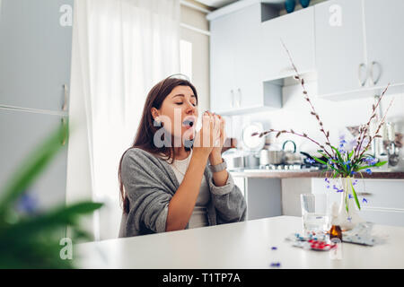Frühling Allergie. Junge Frau Niesen, weil der Blumen mit Pillen auf Küche zu Hause umgeben. Saisonale Allergien Konzept. Stockfoto