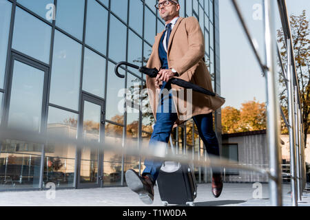 Gut aussehender Geschäftsmann in Gläsern und beige Mantel mit Koffer und Schirm auf der Straße Stockfoto