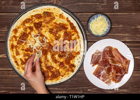 Frau Hand ist das Streuen Pizza mit Käse. Rezept Schritt für Schritt Pizza Capricciosa flatlay auf braunem Holz Stockfoto